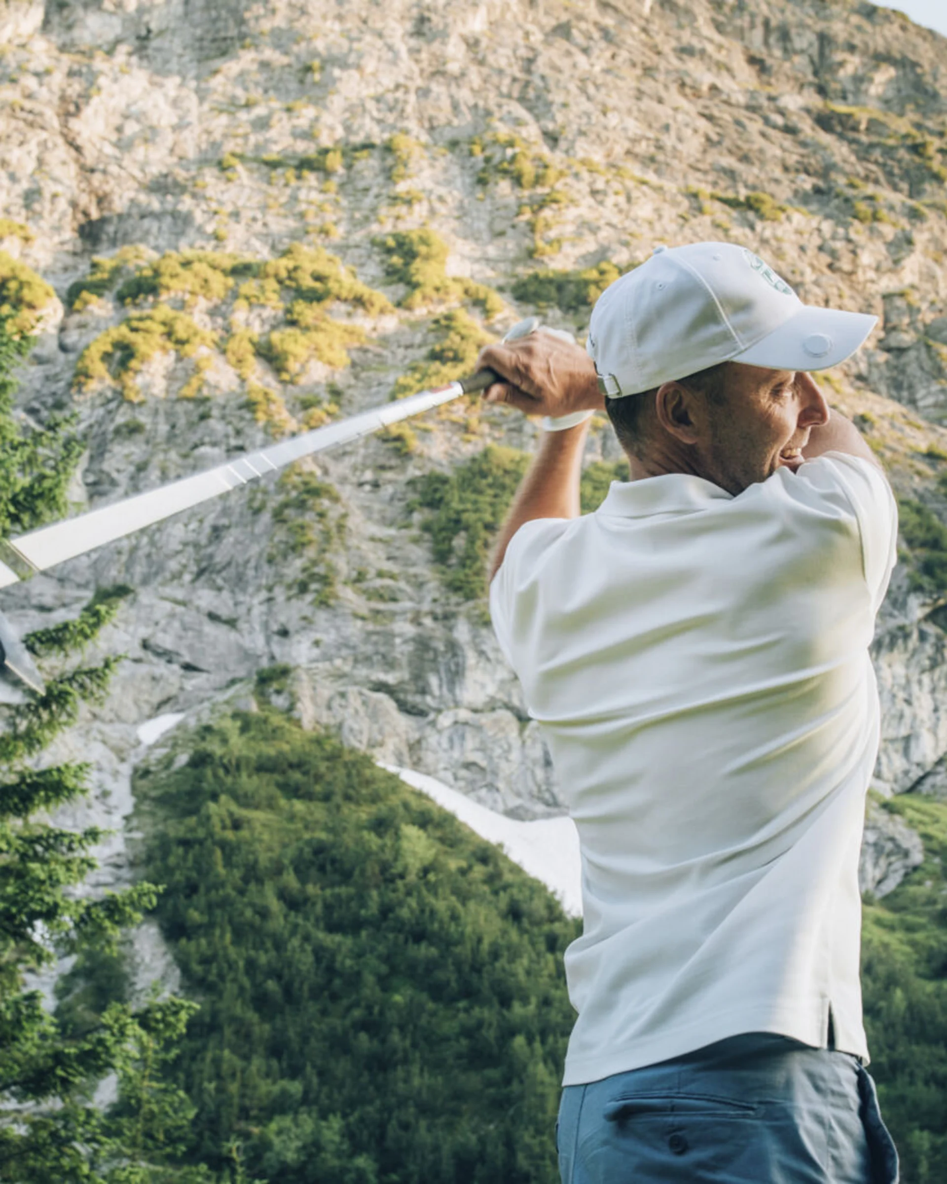 Golfer beim Abschlag vor einer felsigen Bergkulisse und Nadelbäumen