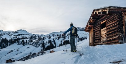 Skitourengeher steht neben einer urigen Hütte und genießt den Ausblick auf die Berge