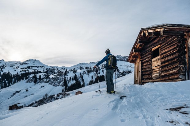 Skitourengeher steht neben einer urigen Hütte und genießt den Ausblick auf die Berge