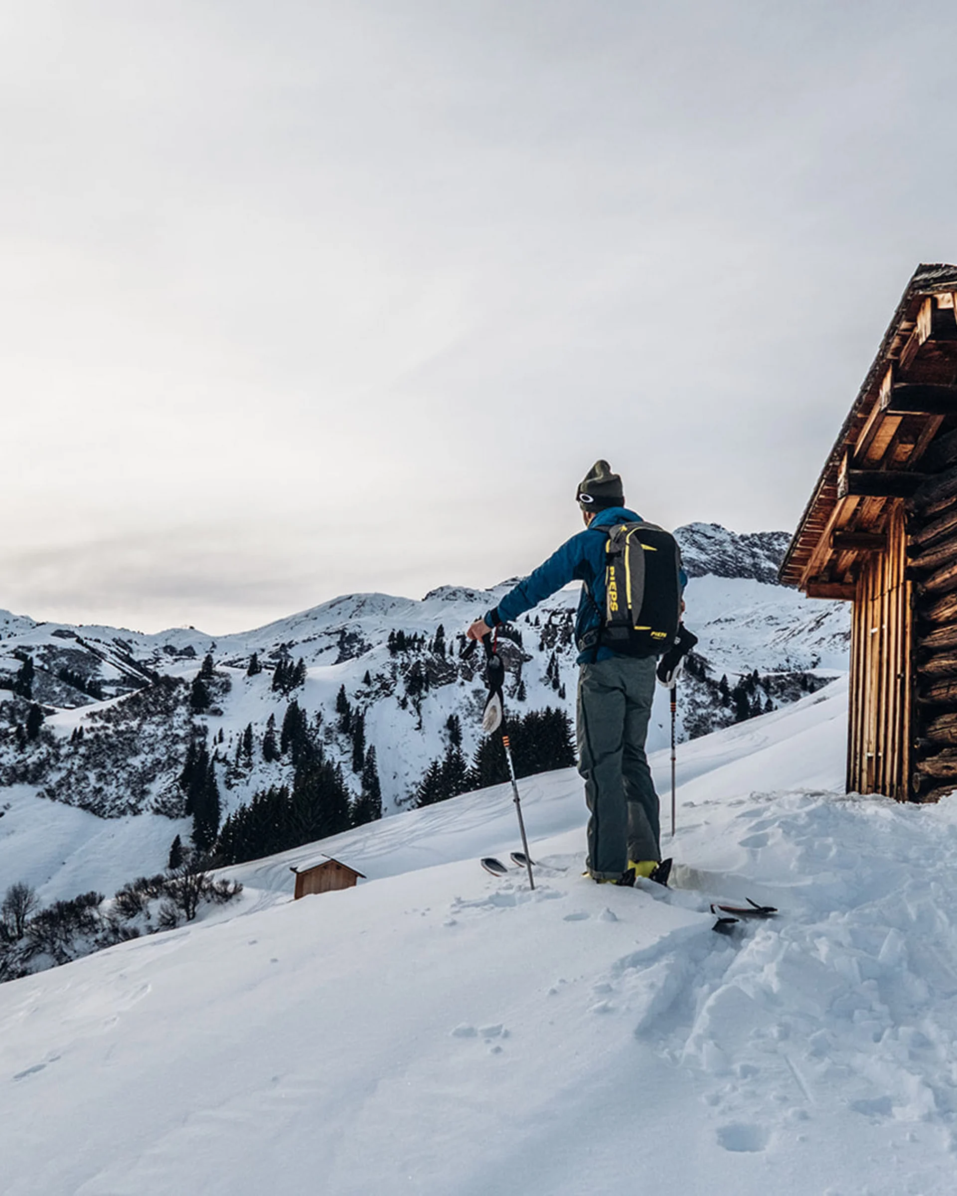 Skifahrer steht neben einer Holzhütte in verschneiter Berglandschaft beim Sonnenuntergang