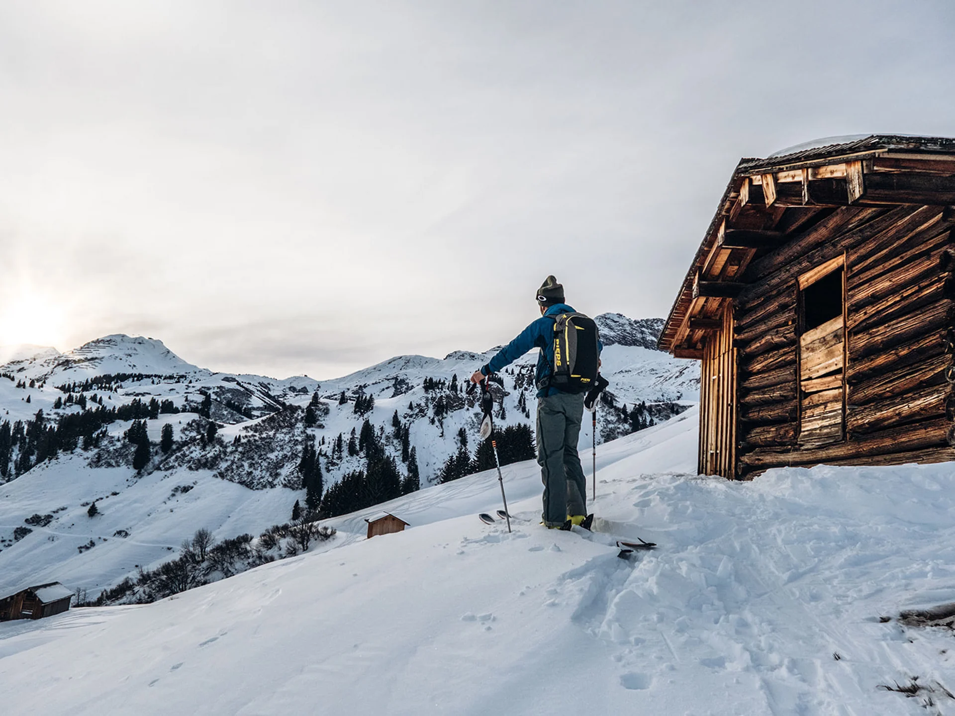 Skier standing by wooden cabin in snowy mountain landscape at sunset