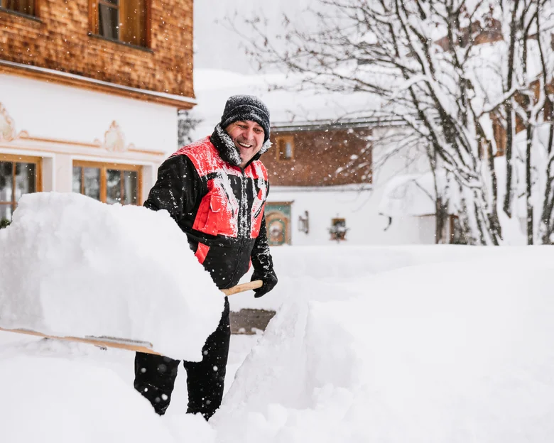 Das Team des Hotel Aurora in Lech Mann in Winterkleidung schaufelt Schnee vor einem verschneiten Haus