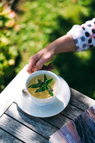 Hand hält Tasse mit Kräutertee auf Holztisch im Garten