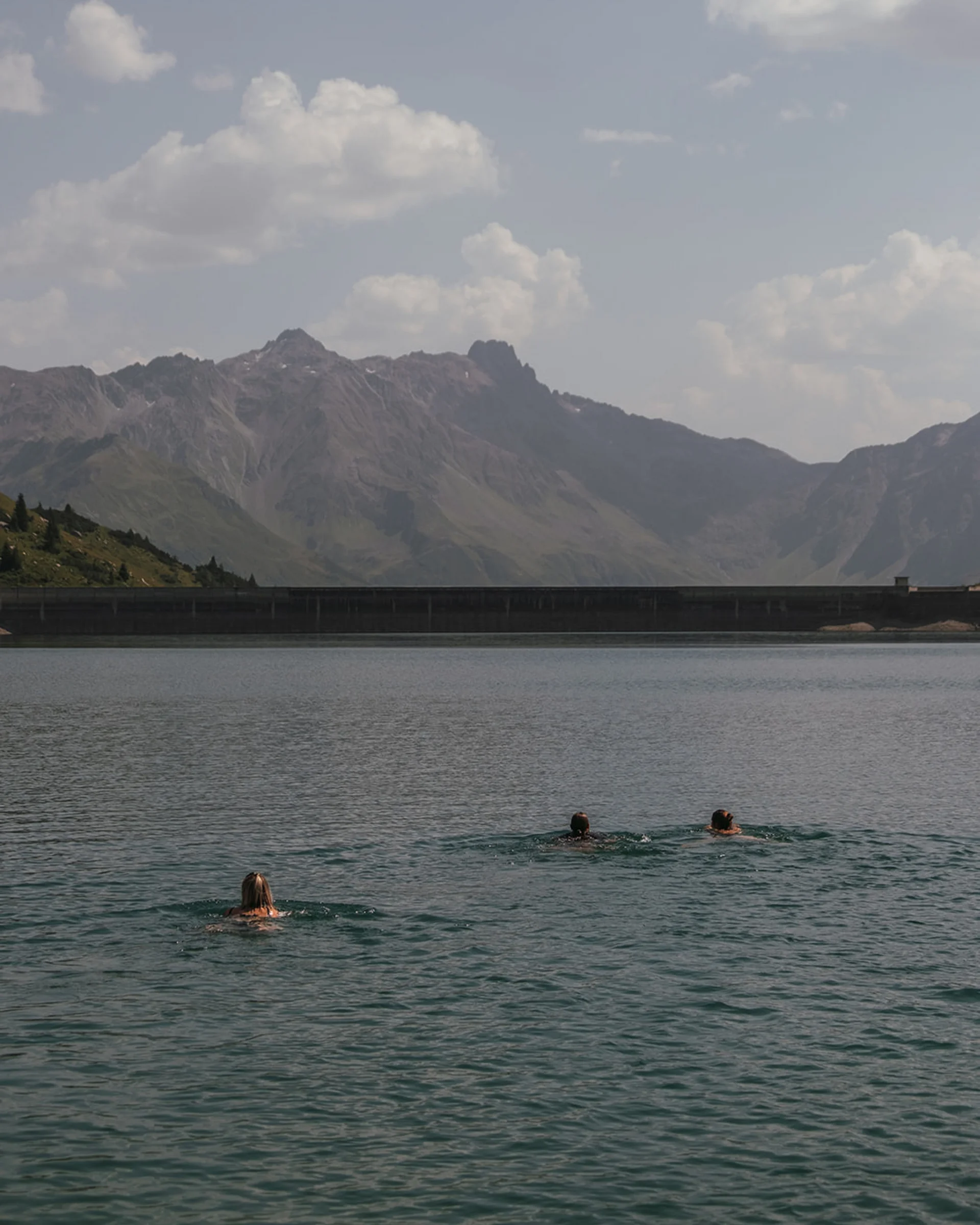 Drei Personen schwimmen in einem See vor einer Berglandschaft bei bewölktem Himmel