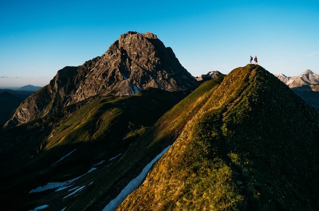 Zwei Personen gehen einen schmalen Pfad entlang und haben ein schönes Bergpanorama um sich. 