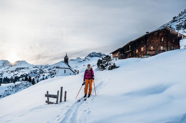 Frau mit Skitourenausrüstung steht in mitten einer Schneelandschaft neben einer urigen Hütte und einer kleinen weißen Kapelle. 