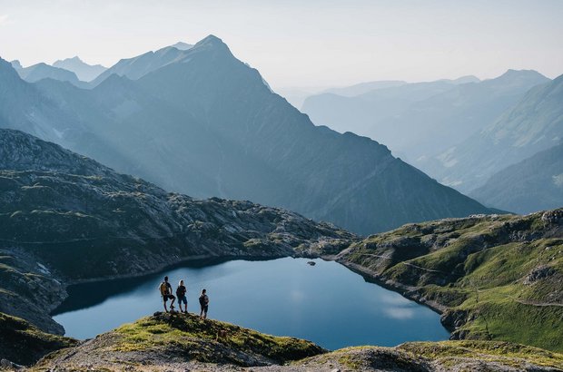 Kleine Wandergruppe genießt den Ausblick auf einen See mit der Berglandschaft im Hintergrund.