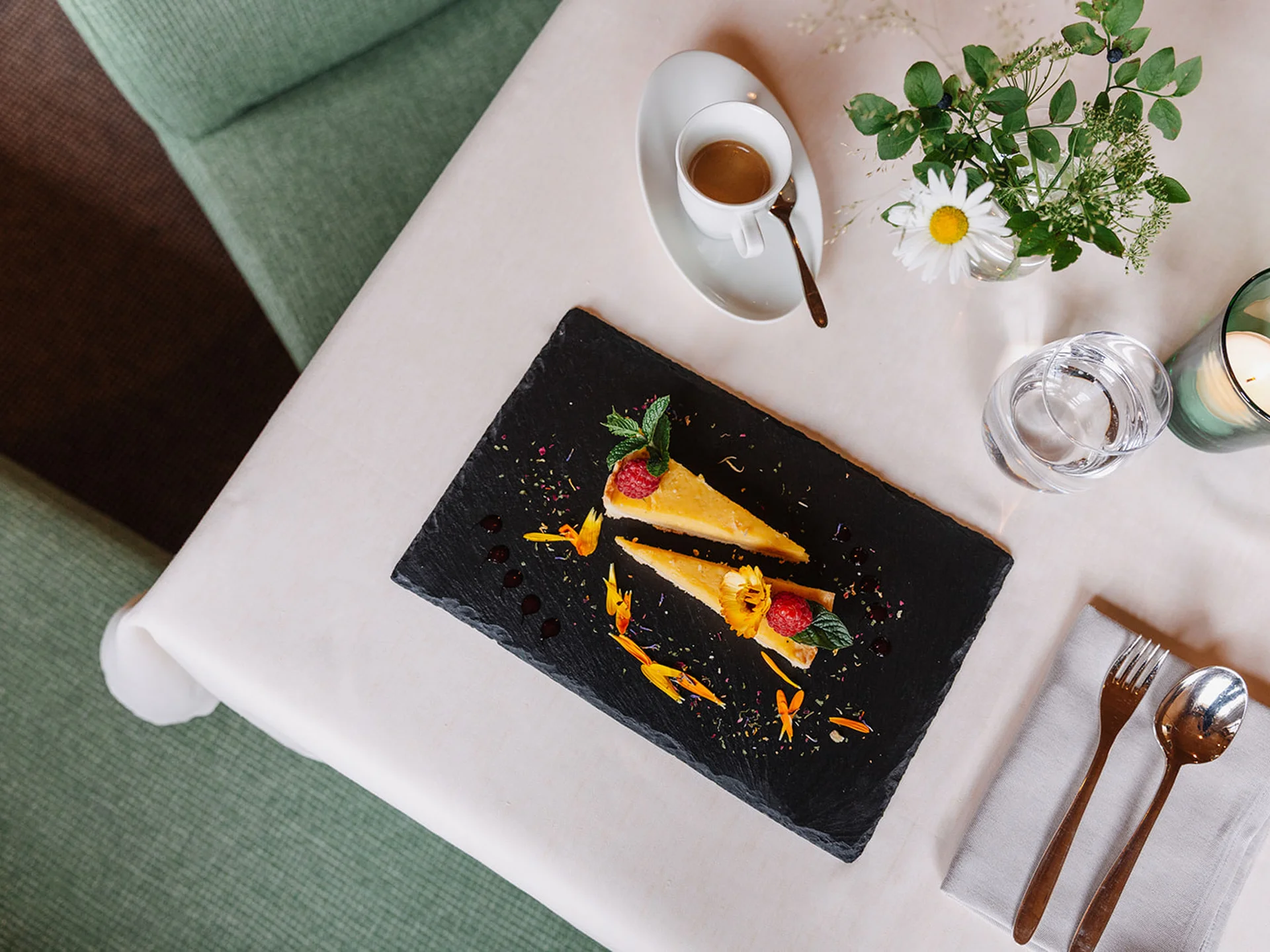 Two cake slices on slate plate with coffee, candle, and flowers on table