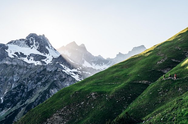 Drei Personen wandern an einem schmalen Pfad entlang mit Ausblick auf die schroffen Gipfel. 