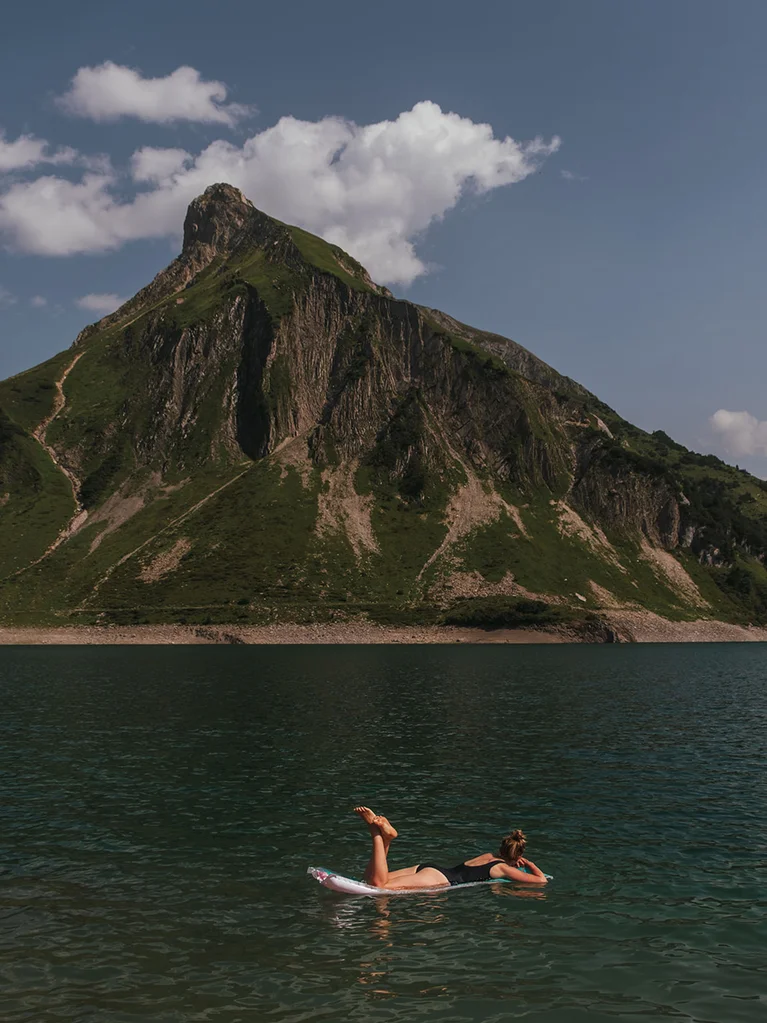 Frau entspannt auf Wasser schwimmend vor Bergkulisse unter bewölktem Himmel