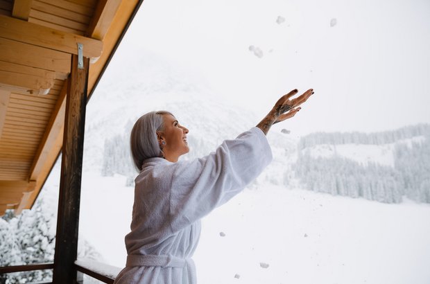 Frau im Bademantel steht am Balkon und spielt mit Schnee. 