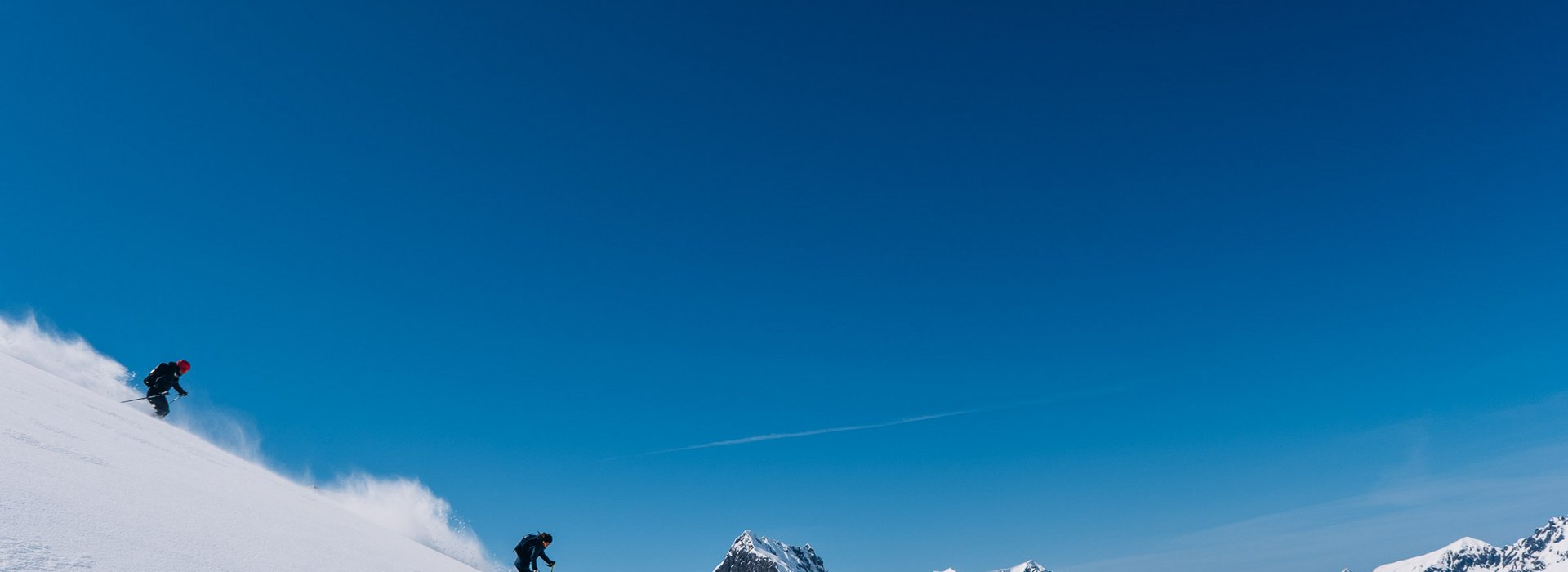 Zwei Menschen fahren durch den Tiefschnee. Im Hintergrund strahlend blauer Himmel und ein wunderschönes Bergpanorama.