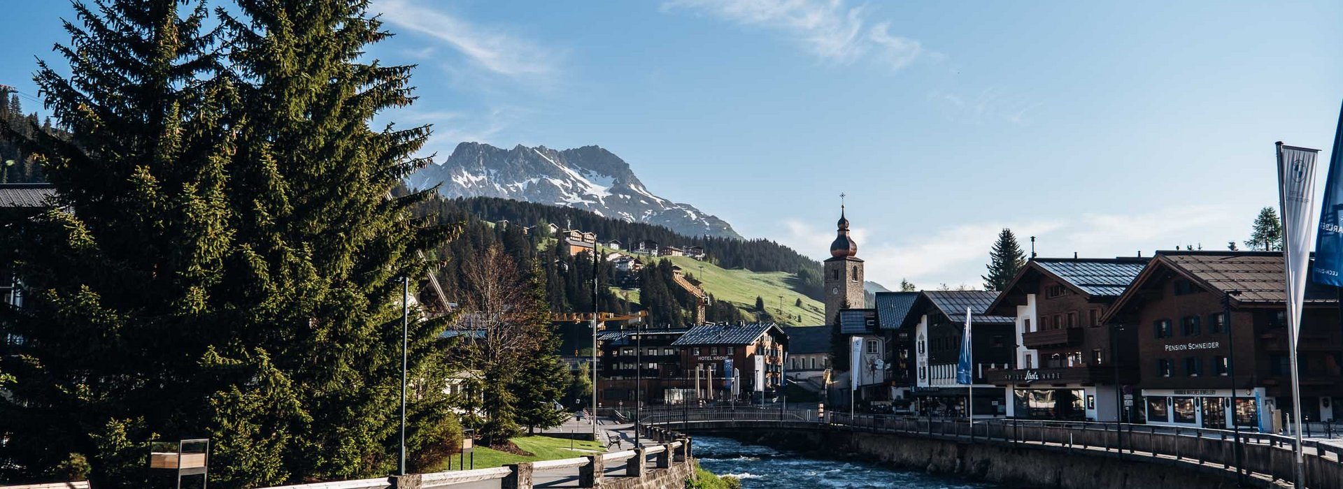 Ausblick auf die Ortschaft Lech mit seinen Häusern und dem Kirchturm. Der Fluss "Lech" der mitten durch fließt und das Bergpanorama im Hintergrund.