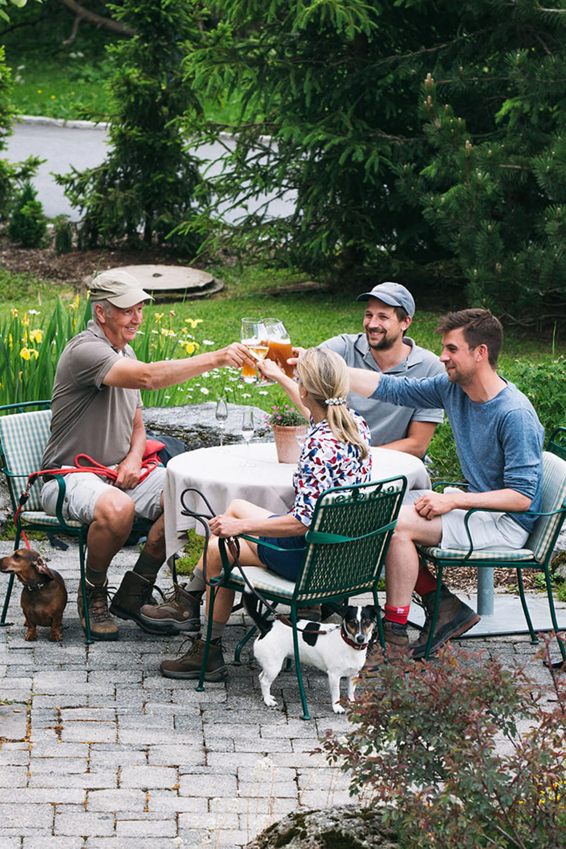 Vier Personen stoßen mit Getränken im Garten an, zwei kleine Hunde sind dabei